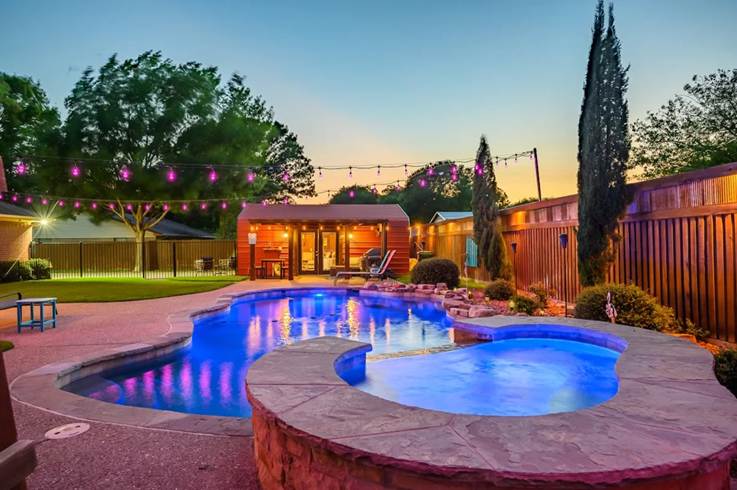 A luxurious backyard oasis at sunset featuring an illuminated custom swimming pool and hot tub, vibrant pink string lights, and a cozy pool house.