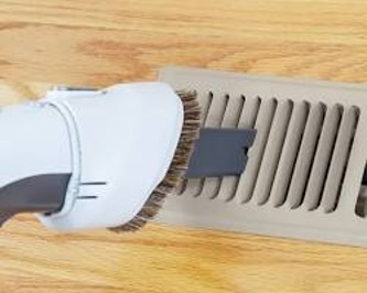 A close-up of a vacuum cleaner's brush attachment clearing dust from a floor vent register on hardwood flooring.