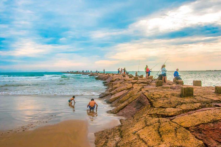 A long rock jetty stretching into the ocean with people fishing and children playing on the adjacent sandy beach under a cloudy blue sky. A Pre-Vacation Home Deep Clean us a must!