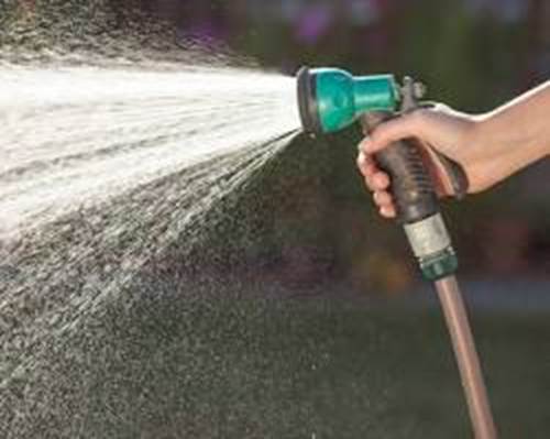 A close-up of a hand holding a garden hose nozzle and spraying a wide mist of water outdoors.
