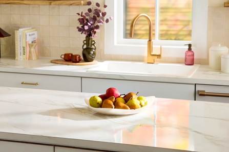 A bright, modern kitchen featuring white marble-look countertops, a bowl of fresh pears on the island, and a gold faucet by the window sink.