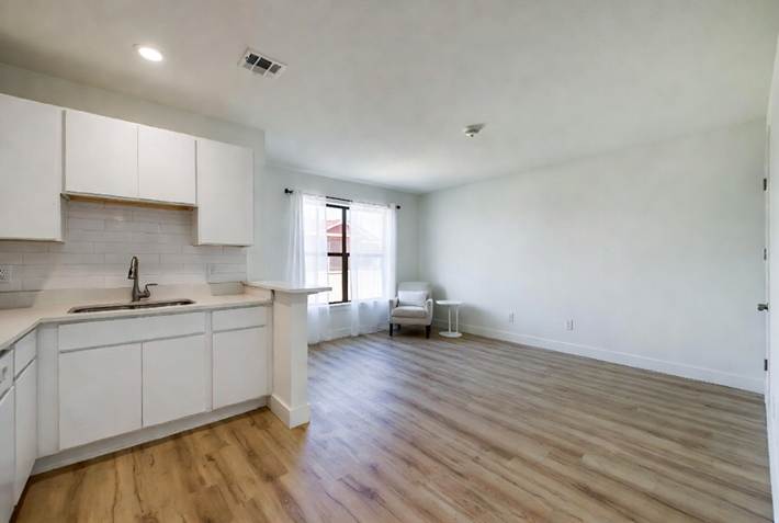 An empty, freshly cleaned open-concept apartment featuring white kitchen cabinets, wood-look flooring, and a bright living area with a window.