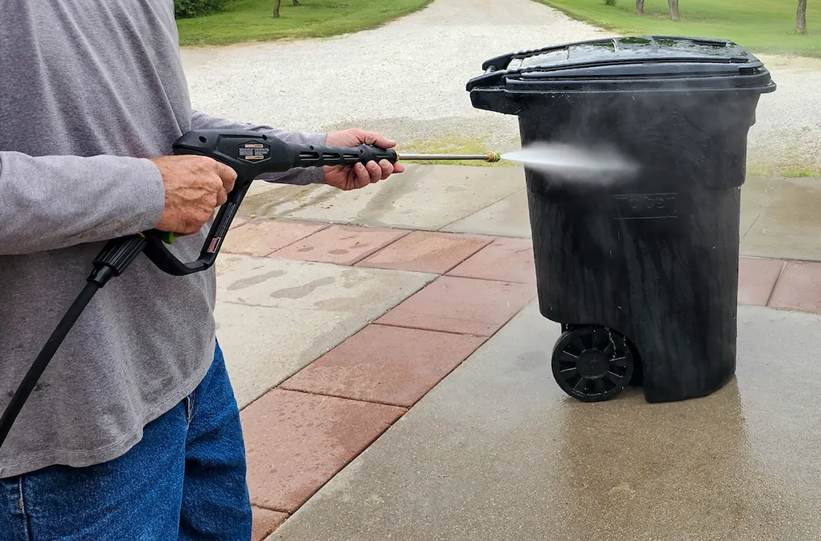 The 15-Minute "Cool Evening" Reset Checklist: A person holding a pressure washer spray wand, directing a high-pressure jet of water to power wash the side of a large black outdoor trash can.