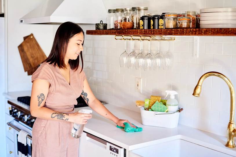 The 15-Minute "Cool Evening" Reset Checklist: A smiling woman with arm tattoos wiping down a white kitchen countertop using a green cloth and a spray bottle in a modern kitchen.