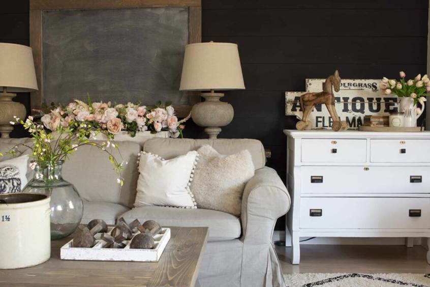 The 15-Minute "Cool Evening" Reset Checklist: A rustic farmhouse-style living room featuring a neutral beige sofa, dark shiplap accent wall, white dresser with vintage decor, and soft floral arrangements.