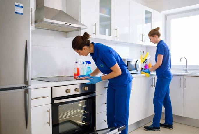 Two professional cleaners wearing blue uniforms and gloves cleaning a modern white kitchen, with one wiping down the stovetop and the other cleaning the counter near the sink.