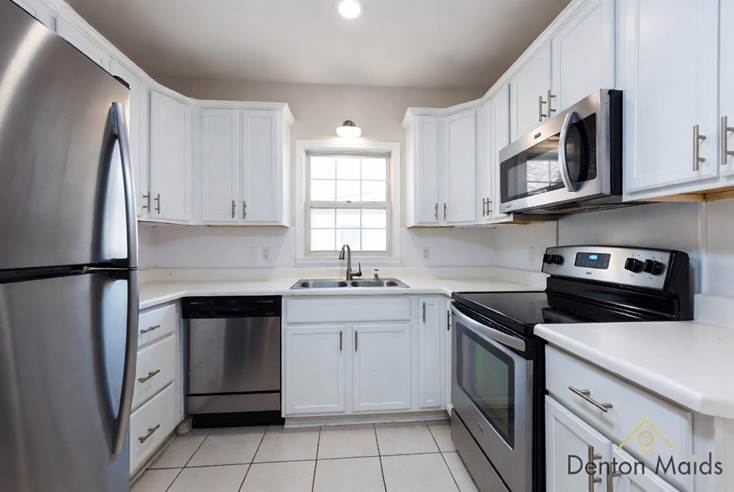 Move-Out Survival Guide: A bright, clean residential kitchen with freshly painted white cabinets, light countertops, and stainless steel appliances, including a large refrigerator on the left and a microwave above an electric range on the right. The floor is tiled, and a central window above the double-basin sink lets in natural light. The Denton Maids logo is in the bottom right corner.