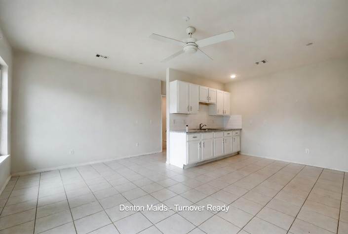 Move-Out Survival Guide: A bright, empty studio apartment or open-concept living space with light-colored tile flooring, beige walls, and a white ceiling fan. A small, clean kitchenette features white upper and lower cabinets, a gray granite countertop, and a stainless steel sink. Text overlay at the bottom reads "Denton Maids - Turnover Ready."