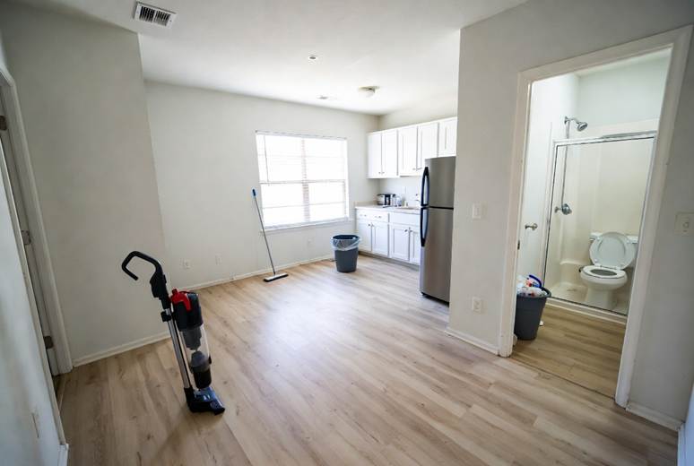 An empty, clean apartment interior with light wood-plank flooring. In the foreground, a black and red upright vacuum cleaner stands ready. In the background, there is a small kitchenette with white cabinets and a stainless steel refrigerator, next to a window. An open doorway on the right reveals a tidy bathroom with a toilet and a glass-enclosed shower.