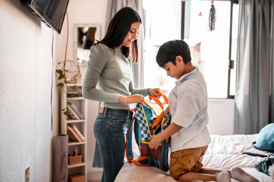 Back-to-School Prep: A smiling mother helping her young son pack a colorful blue and orange backpack on a bed in a bright bedroom.