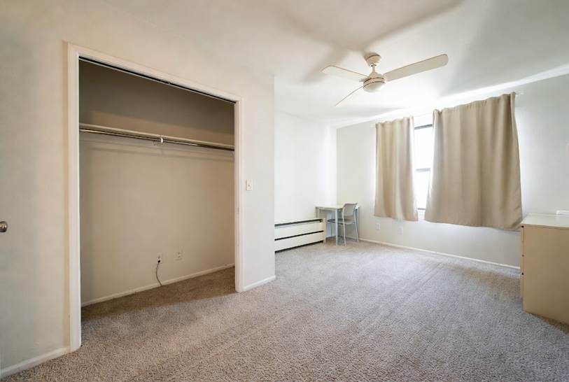 The Ultimate Denton Student Move-In & Move-Out Cleaning Checklist: Spacious, empty bedroom featuring neutral beige carpet, white walls, an open closet with a hanging rod, a ceiling fan, beige curtains over a window, and a small desk setup in the corner.