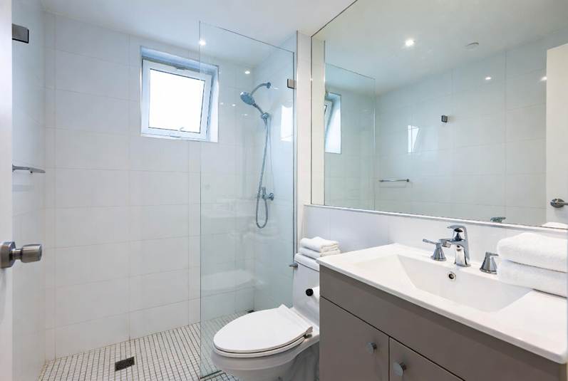 The Ultimate Denton Student Move-In & Move-Out Cleaning Checklist: Bright, modern bathroom featuring white wall tiles, a walk-in shower with a glass divider, a large mirror over a vanity with a white basin and grey cabinet, a toilet, and neatly folded white towels.