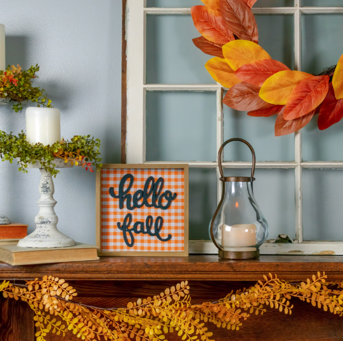 Autumn mantel display featuring a "hello fall" orange gingham sign, a glass candle lantern, and a fall leaf wreath against a white window frame.
