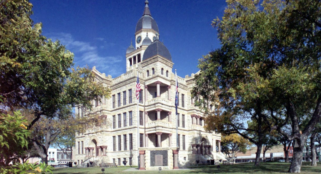 North Texas Fall Prep: Historic stone courthouse with a central clock tower and dome, framed by lush green trees under a clear blue sky.