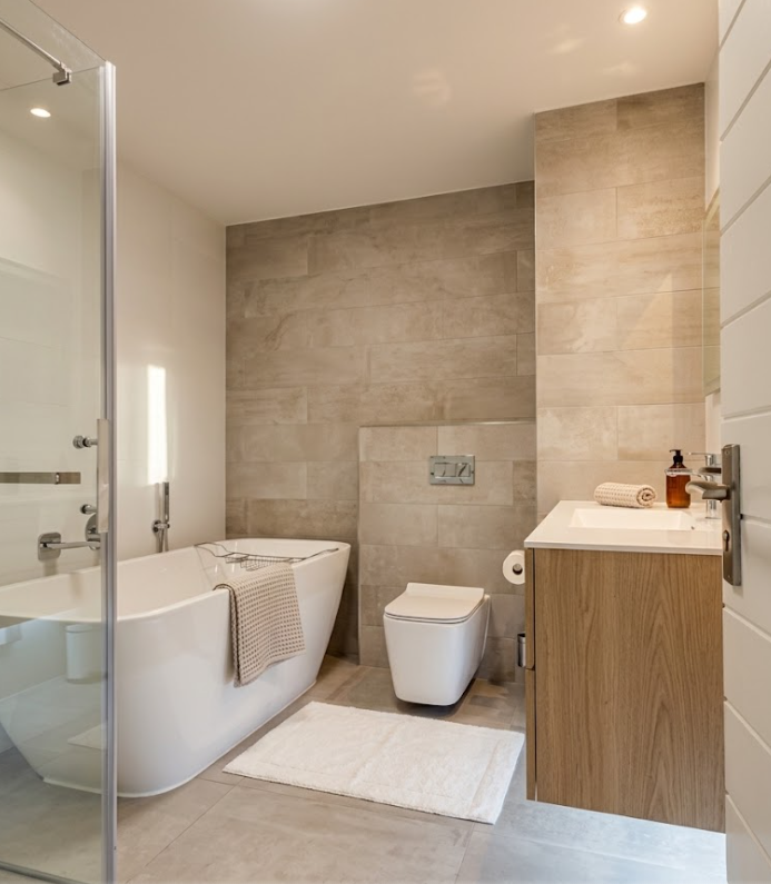 Modern, upscale bathroom featuring a freestanding soaking tub behind a glass enclosure, warm wood floating vanity, wall-hung toilet, and natural stone-look tiles.
