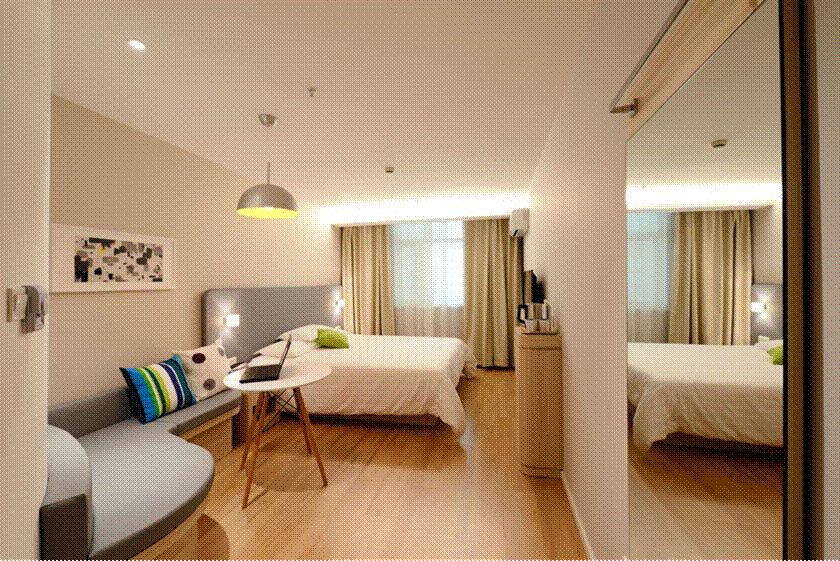 Guest Room Ready! Modern, brightly lit hotel-style guest bedroom featuring a neatly made bed with white linens, curved grey seating nook, work table with laptop, and large wall mirror.