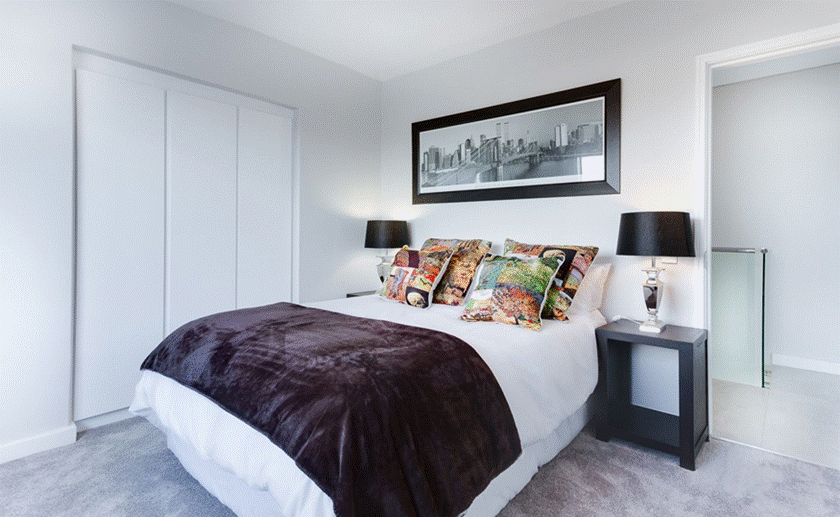 Guest Room Ready: A cozy, modern bedroom with a neatly made white bed, dark faux-fur throw blanket, colorful patterned pillows, matching black bedside lamps, and a framed city skyline print on the wall