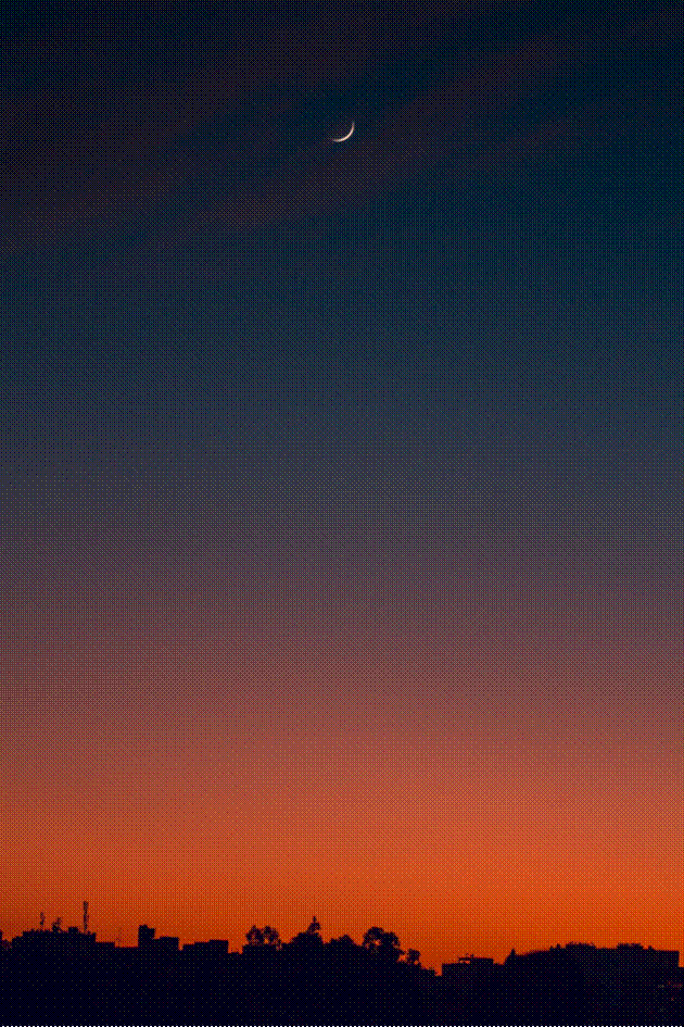 Slim crescent moon shining in a twilight sky transitioning from fiery orange to deep blue over a dark city skyline silhouette.