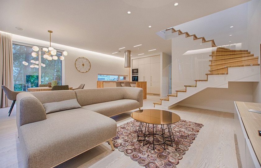 Modern open-concept living room with a beige sectional sofa, circular coffee table, pattern rug, and a floating wooden staircase with glass railings.